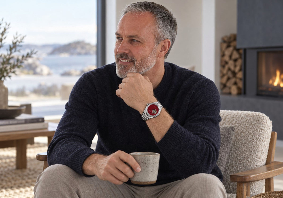 Man holding a ceramic mug, wearing a SANDAHL ACHROMATIC watch with a metallic red open-heart dial and stainless steel bracelet.
