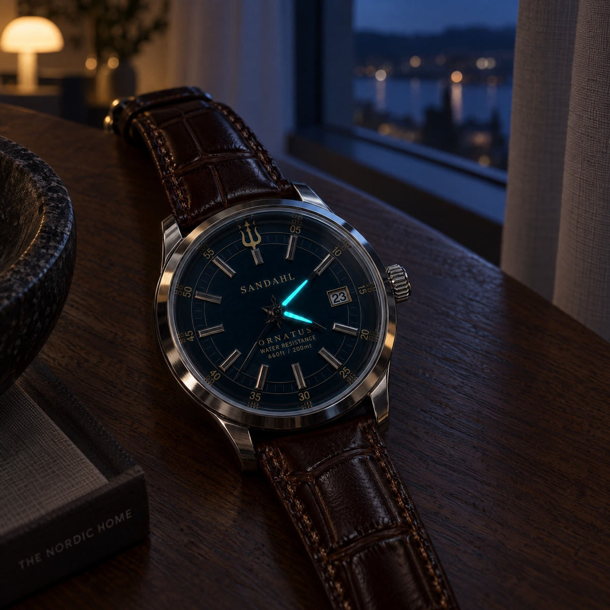 SANDAHL ORNATUS watch with glowing hands and a brown leather strap on a wooden surface beside a window at dusk.
