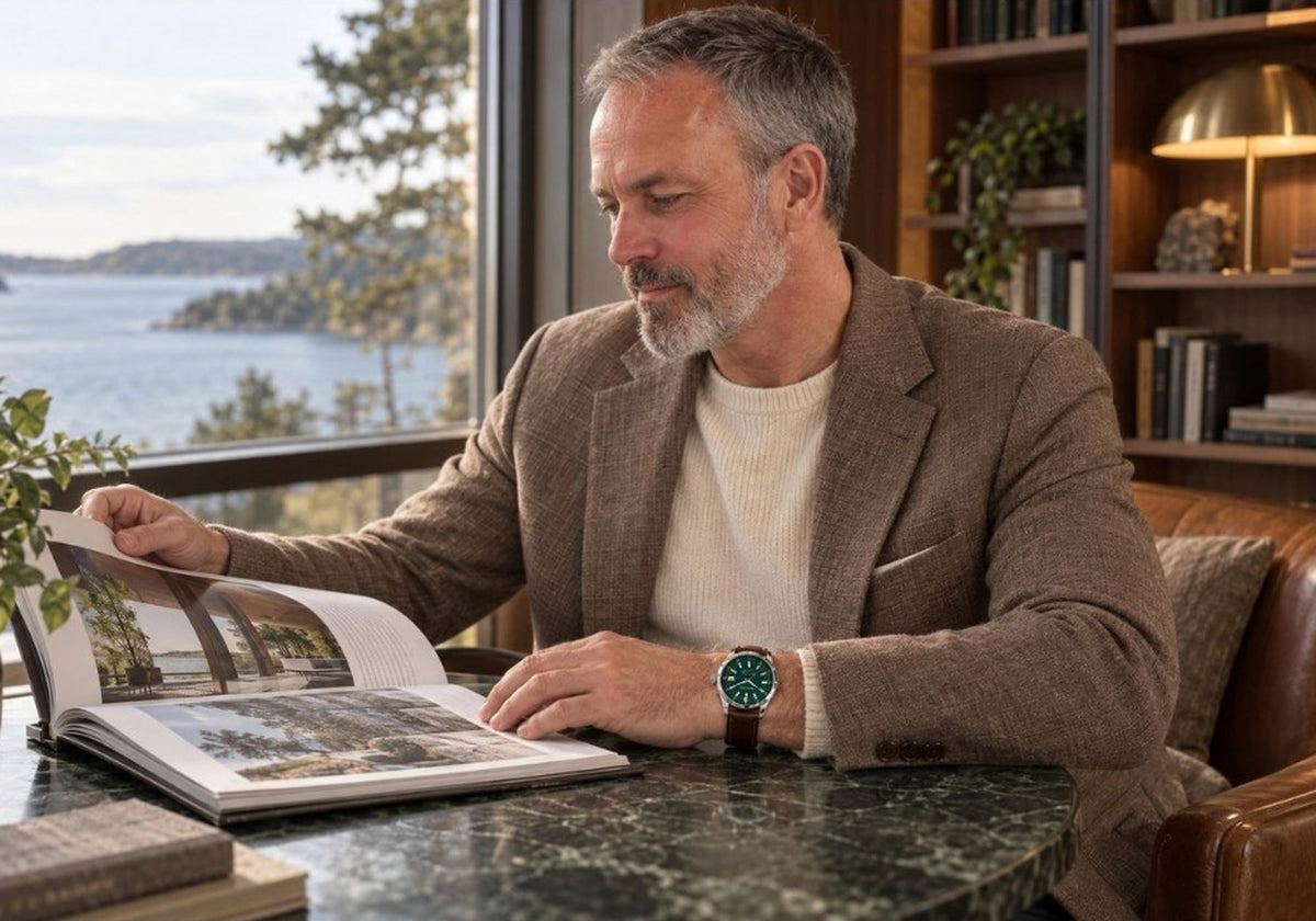 Man wearing a SANDAHL ORNATUS Malachite watch with a green dial while reading a book beside a window overlooking the water.