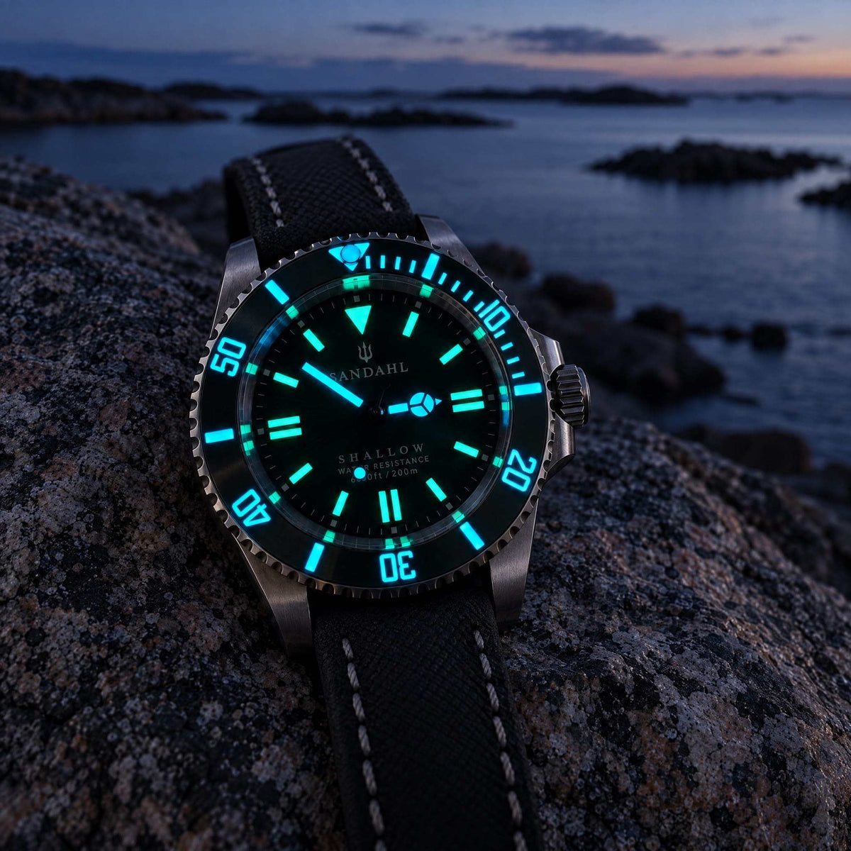 SANDAHL SHALLOW watch with glowing hands, hour markers and bezel markings on coastal granite at dusk.
