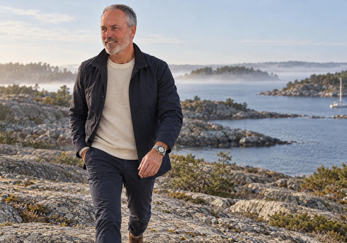Man walking along coastal rocks wearing a SANDAHL SHALLOW Sea Mist Silver watch on his left wrist, with misty islands behind him.