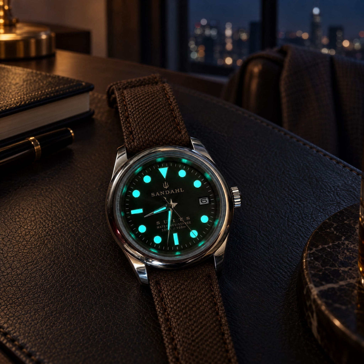 SANDAHL SUITS watch with glowing hands and hour markers on a dark desk beside a notebook, with city lights in the background.