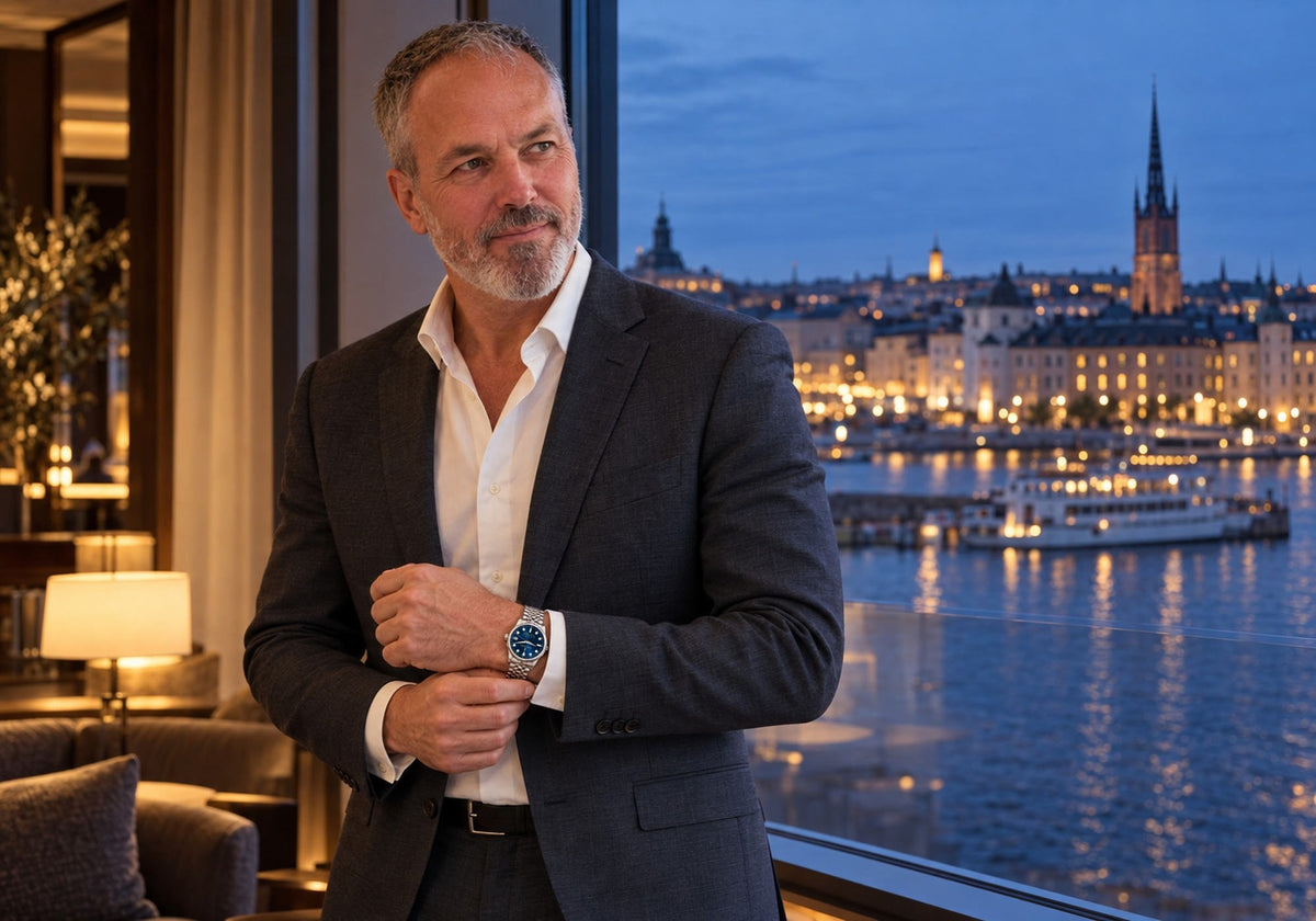 Man in a charcoal suit adjusting his cuff, wearing a SANDAHL SUITS Midnight Navy watch beside a window overlooking Stockholm’s waterfront at dusk.