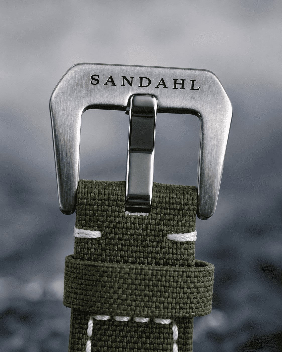 Close-up of the SANDAHL-engraved steel buckle and olive-green textile strap for SUITS.