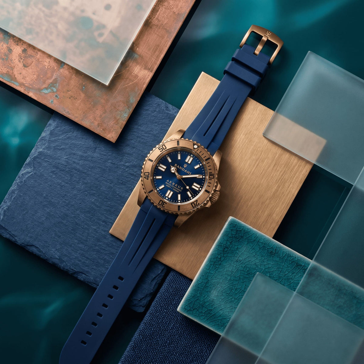 SANDAHL AEREUS Abyss Blue bronze watch with a blue rubber strap, arranged among blue, green and bronze-toned materials.