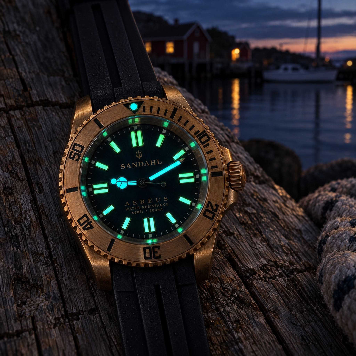 SANDAHL AEREUS bronze watch with glowing hands and hour markers on weathered wood beside a harbour at dusk.