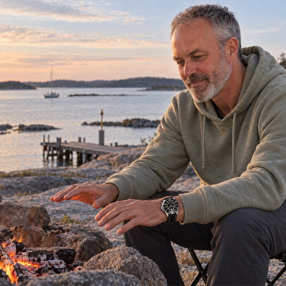 Man wearing a SANDAHL CENTURION Blackwater watch while warming his hands by a shoreline campfire in the Swedish archipelago.