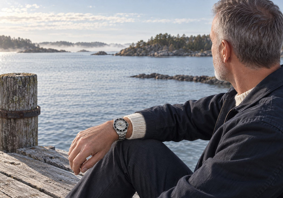 Man wearing a SANDAHL SHALLOW Sea Mist Silver watch with a steel bracelet, seated by the water in the Swedish archipelago.