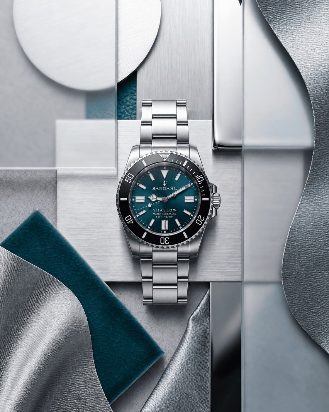 SANDAHL SHALLOW Skagerrak Teal with a steel bracelet, arranged among teal, grey and white material samples.