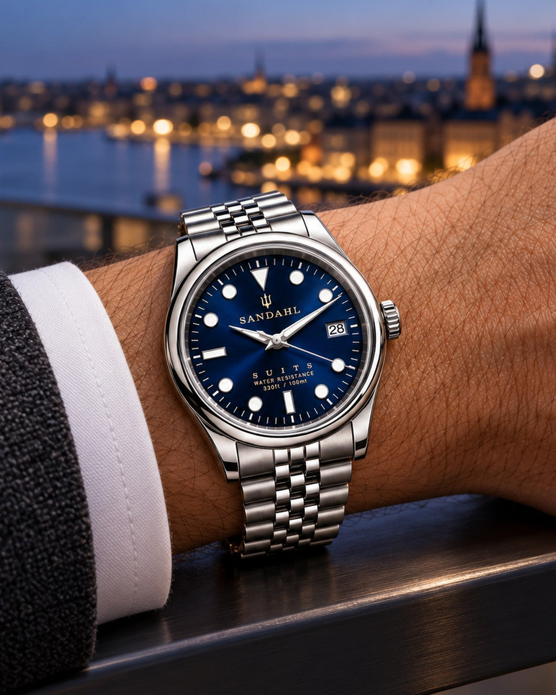 SANDAHL SUITS Midnight Navy with a blue sunburst dial and Jubilee bracelet, worn on the left wrist with a charcoal suit overlooking city lights at dusk.
