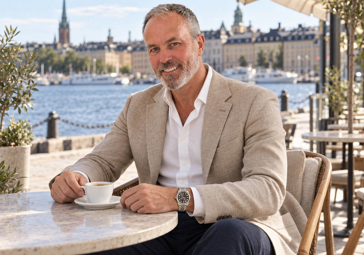 Man in a beige linen blazer wearing a SANDAHL SUITS Savile Taupe watch with a stainless steel Jubilee bracelet, enjoying espresso on a waterfront terrace.